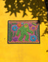 A rectangular flat-printed doormat featuring a faux-tufted, trompe-l'œil texture design. The background is a vibrant hot pink, bordered by a wavy yellow line. In the center is a bright green, smiling Bigfoot or Sasquatch figure with long shaggy hair. Surrounding the cryptid are highly detailed, colorful faux-embroidered floral motifs including blue, orange, and purple daisies, alongside green fern fronds and leaves. The mat is finished with a solid black fabric trim around the outer edge.