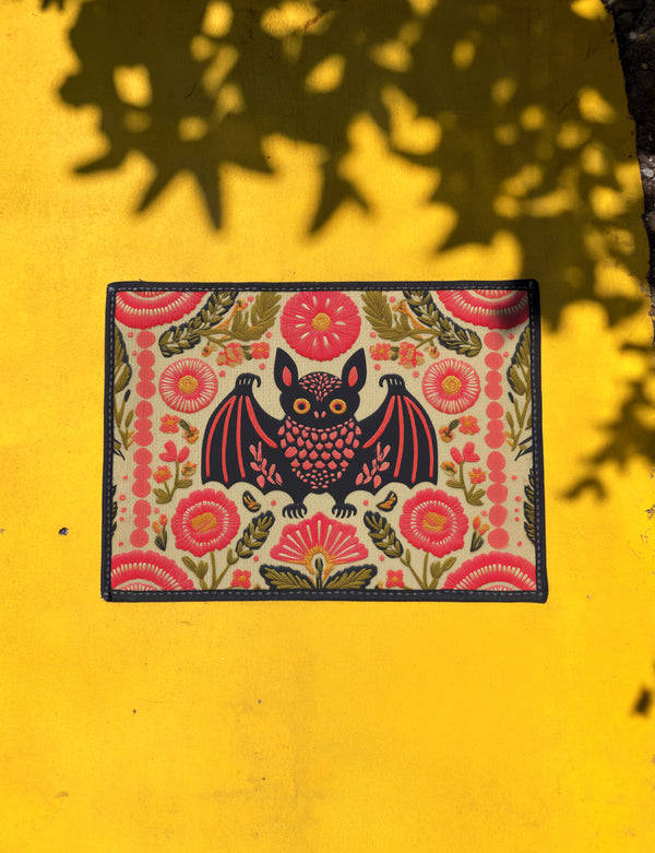 A rectangular beige doormat featuring a large, symmetrical folk art bat illustration in the center. The bat is black with coral red and mustard gold details that mimic the look of satin stitch embroidery. Surrounding the bat are stylized floral motifs in deep pink, coral, and olive green, arranged in a dense, decorative pattern. The texture visually resembles woven linen but is a smooth, flat print. The mat has a solid black trim border and is shown on a flat surface.