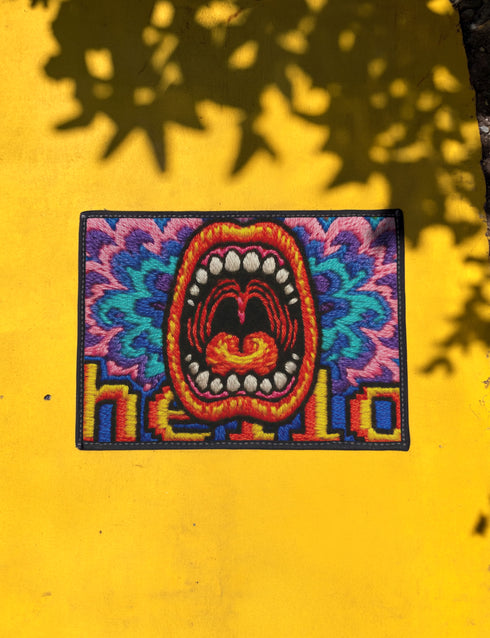 A rectangular indoor and outdoor doormat featuring a vibrant, psychedelic design. The central graphic is a large, open screaming red and orange mouth with white teeth, surrounded by highly saturated wavy, trippy patterns in blue, purple, magenta, and teal. Yellow stylized text reading HELLO is integrated into the pattern. The mat has a smooth 2D flat print surface that mimics the look of textured chainstitch embroidery, finished with a solid black fabric trim around all four outer edges.