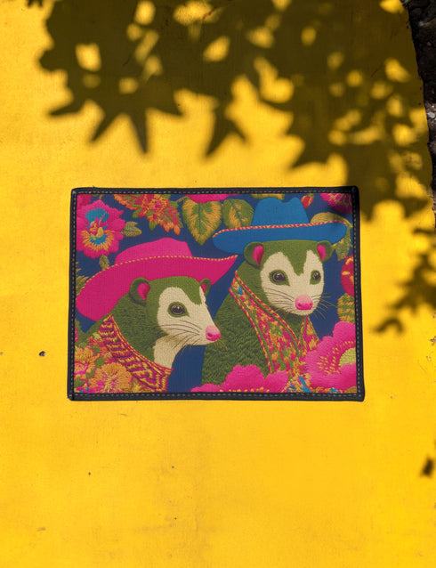 A rectangular non-slip doormat with a black trimmed edge. The mat features a vibrant, colorful folk art illustration on a dark navy blue background with a faux embroidered texture. The design depicts two friendly opossums, rendered in an olive green color with white faces and pink noses. They are surrounded by large, stylized flowers and leaves in shades of hot pink, red, turquoise, and yellow, creating a busy, maximalist pattern. The overall aesthetic is eclectic, whimsical, & highly detailed.