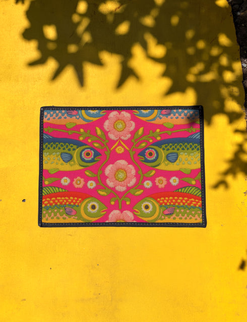 Rectangular indoor outdoor doormat featuring a vibrant hot pink background with a symmetrical maximalist pattern. The design showcases stylized sardines in yellow, lime green, and cobalt blue swimming horizontally towards a central vertical axis of blooming pale pink flowers and green vines. The imagery mimics the texture of vintage needlepoint embroidery but is a flat print on polyester. The mat is framed by a sleek black trim and features a non-slip rubber backing for safety.