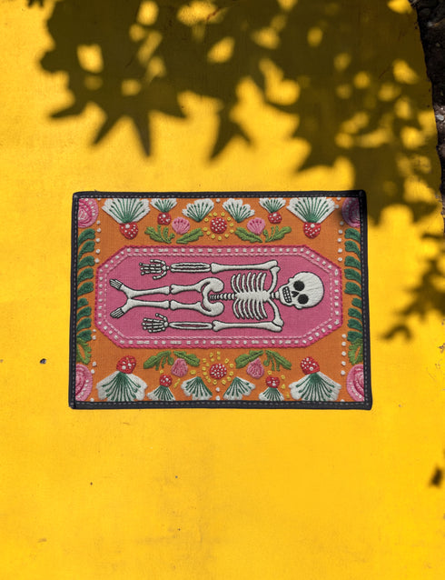 A rectangular terracotta orange doormat featuring a central illustration of a white skeleton lying inside a pink coffin shape. The design is surrounded by a symmetrical pattern of green leaves, pink flowers, and red and white spotted mushrooms. The imagery uses a trompe-l'œil effect to mimic the texture of raised needlepoint embroidery, but the surface is flat printed polyester. The mat is framed with a solid black edge.
