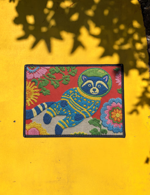 A rectangular outdoor doormat featuring a maximalist 2D print with a Trompe-l'œil embroidery effect. The central design depicts a blue and gold patterned raccoon wearing a green astronaut space helmet. The background is a vibrant red, filled with large, multi-colored folk art flowers in shades of pink, magenta, and gold with green leaves. The mat is finished with a sleek black border and has a textured surface mimicking high-fidelity raised needlepoint stitching.
