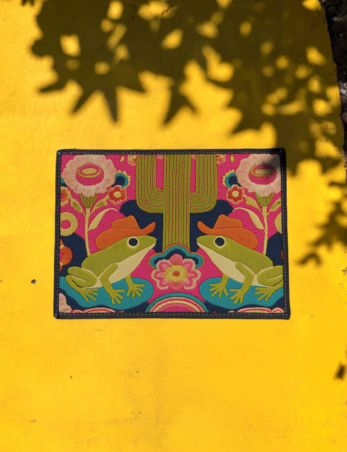 A rectangular polyester doormat with a black trim and non-slip rubber backing. The vibrant, colorful design features a symmetrical illustration on a dark navy blue background. In the center, two green frogs wearing orange cowboy hats sit facing each other. Between them is a tall, olive-green saguaro cactus. The scene is filled with large, psychedelic-style flowers in hot pink, orange, and yellow. The overall aesthetic is funky, retro 70s, maximalist, and trippy, mimicking the look of embroidery.