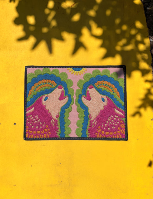 A rectangular outdoor doormat featuring a symmetrical graphic illustration of two howling wolves facing upward in profile. The design utilizes a vibrant psychedelic folk art style with a color palette of magenta pink wolves, peach muzzles, and scalloped layers of lime green and cyan blue foliage. A marigold yellow sun sits at the top center. The background is a dusty rose pink. The texture is a smooth, durable flat 2D print on polyester with a black non-slip rubber border.