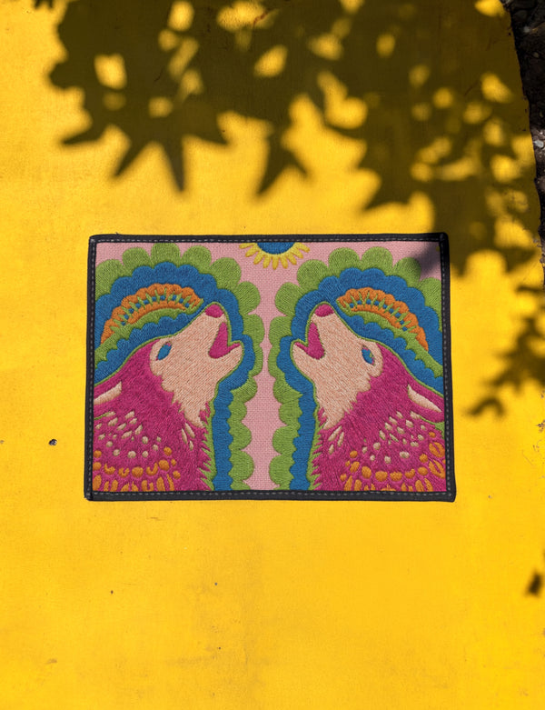 A rectangular outdoor doormat featuring a symmetrical graphic illustration of two howling wolves facing upward in profile. The design utilizes a vibrant psychedelic folk art style with a color palette of magenta pink wolves, peach muzzles, and scalloped layers of lime green and cyan blue foliage. A marigold yellow sun sits at the top center. The background is a dusty rose pink. The texture is a smooth, durable flat 2D print on polyester with a black non-slip rubber border.