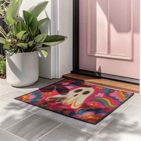 A rectangular polyester doormat with a vibrant, psychedelic design on a hot pink background. In the center is a cute, cartoonish ghost in off-white with yellow and orange outlines; it has large, dark, expressive eyes. The ghost is surrounded by a maximalist pattern of 70s-style floral motifs and puffy clouds in shades of teal, blue, orange, and light pink. Two small, colorful rainbows arc over the ghost's shoulders. The doormat has a durable, textured surface and is framed by a sleek black trim.