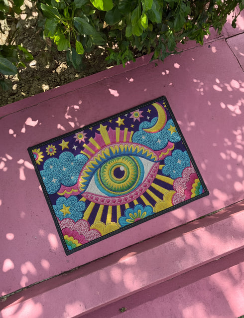Rectangular entryway doormat with a deep purple background printed in a 2D faux embroidery illusion. The symmetrical mandala centers on a large green, pink, and white evil eye with golden yellow sunburst lines radiating outward. The corners hold textured cyan clouds, yellow crescent moons, and stars, with a scalloped floral motif along the lower edge. Finished with black trim and a non-slip rubber backing. Psychedelic celestial maximalist home decor and protective evil eye welcome mat for bold, eclectic ent