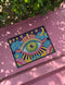 Rectangular entryway doormat with a deep purple background printed in a 2D faux embroidery illusion. The symmetrical mandala centers on a large green, pink, and white evil eye with golden yellow sunburst lines radiating outward. The corners hold textured cyan clouds, yellow crescent moons, and stars, with a scalloped floral motif along the lower edge. Finished with black trim and a non-slip rubber backing. Psychedelic celestial maximalist home decor and protective evil eye welcome mat for bold, eclectic ent
