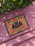 A rectangular beige doormat featuring a large, symmetrical folk art bat illustration in the center. The bat is black with coral red and mustard gold details that mimic the look of satin stitch embroidery. Surrounding the bat are stylized floral motifs in deep pink, coral, and olive green, arranged in a dense, decorative pattern. The texture visually resembles woven linen but is a smooth, flat print. The mat has a solid black trim border and is shown on a flat surface.
