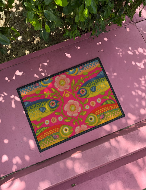 A rectangular doormat featuring a large hot pink smiley face on a deep teal background. The smiley face has green cannabis leaves for eyes and a yellow stitched-style smile. The central face is surrounded by stylized green foliage arches and concentric circles in pink, yellow, and white, resembling abstract flowers. The design mimics a vintage needlepoint or tapestry texture but is a flat print. The mat has a solid black trim border.