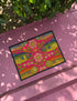 A rectangular doormat featuring a large hot pink smiley face on a deep teal background. The smiley face has green cannabis leaves for eyes and a yellow stitched-style smile. The central face is surrounded by stylized green foliage arches and concentric circles in pink, yellow, and white, resembling abstract flowers. The design mimics a vintage needlepoint or tapestry texture but is a flat print. The mat has a solid black trim border.