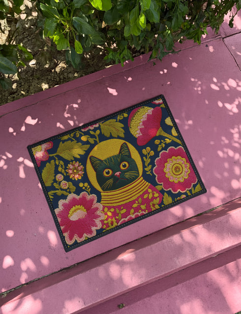 High-fidelity printed doormat featuring a "Space Kitty" design in a tapestry-style faux-texture. A black cat with large yellow eyes wears a golden ribbed space helmet, centered on a deep navy background. The cat is surrounded by oversized, vibrant magenta and pink folk-art flowers with chartreuse and olive green leaves. The design uses a printed illusion to mimic 3D embroidery stitching. The mat is rectangular with a sleek black border and a non-slip backing.