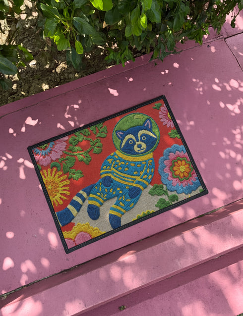 A rectangular outdoor doormat featuring a maximalist 2D print with a Trompe-l'œil embroidery effect. The central design depicts a blue and gold patterned raccoon wearing a green astronaut space helmet. The background is a vibrant red, filled with large, multi-colored folk art flowers in shades of pink, magenta, and gold with green leaves. The mat is finished with a sleek black border and has a textured surface mimicking high-fidelity raised needlepoint stitching.