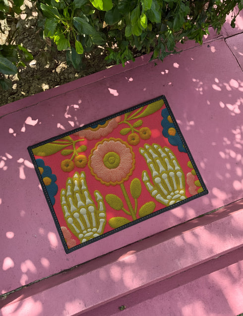 Pink outdoor doormat featuring a symmetrical folk art design. Two white skeleton hands with palms facing forward frame a large central pink and peach flower with a yellow center. Olive green stems, gold berries, and teal blue floral accents decorate the vibrant magenta background. The design mimics a textured embroidery look but is a flat 2d print. Framed with a sleek black border.