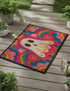 A rectangular polyester doormat with a vibrant, psychedelic design on a hot pink background. In the center is a cute, cartoonish ghost in off-white with yellow and orange outlines; it has large, dark, expressive eyes. The ghost is surrounded by a maximalist pattern of 70s-style floral motifs and puffy clouds in shades of teal, blue, orange, and light pink. Two small, colorful rainbows arc over the ghost's shoulders. The doormat has a durable, textured surface and is framed by a sleek black trim.