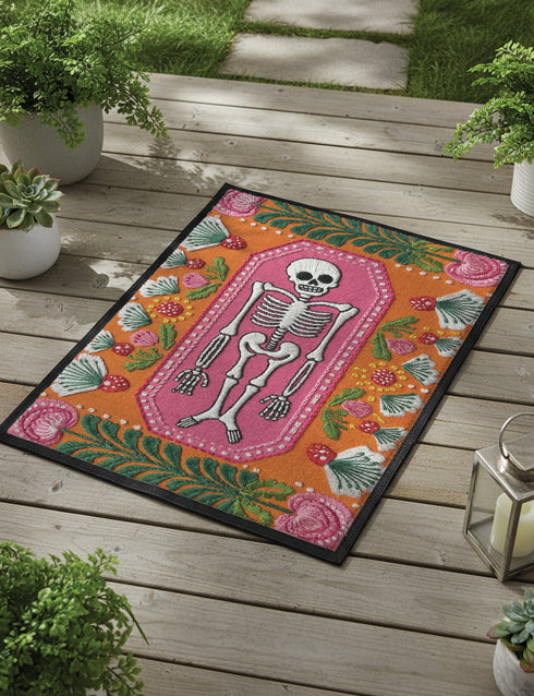 A rectangular terracotta orange doormat featuring a central illustration of a white skeleton lying inside a pink coffin shape. The design is surrounded by a symmetrical pattern of green leaves, pink flowers, and red and white spotted mushrooms. The imagery uses a trompe-l'œil effect to mimic the texture of raised needlepoint embroidery, but the surface is flat printed polyester. The mat is framed with a solid black edge.