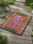A rectangular terracotta orange doormat featuring a central illustration of a white skeleton lying inside a pink coffin shape. The design is surrounded by a symmetrical pattern of green leaves, pink flowers, and red and white spotted mushrooms. The imagery uses a trompe-l'œil effect to mimic the texture of raised needlepoint embroidery, but the surface is flat printed polyester. The mat is framed with a solid black edge.