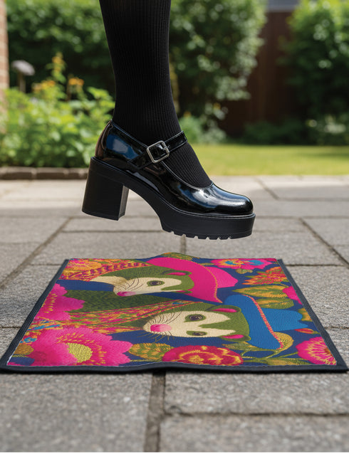 A rectangular non-slip doormat with a black trimmed edge. The mat features a vibrant, colorful folk art illustration on a dark navy blue background with a faux embroidered texture. The design depicts two friendly opossums, rendered in an olive green color with white faces and pink noses. They are surrounded by large, stylized flowers and leaves in shades of hot pink, red, turquoise, and yellow, creating a busy, maximalist pattern. The overall aesthetic is eclectic, whimsical, & highly detailed.