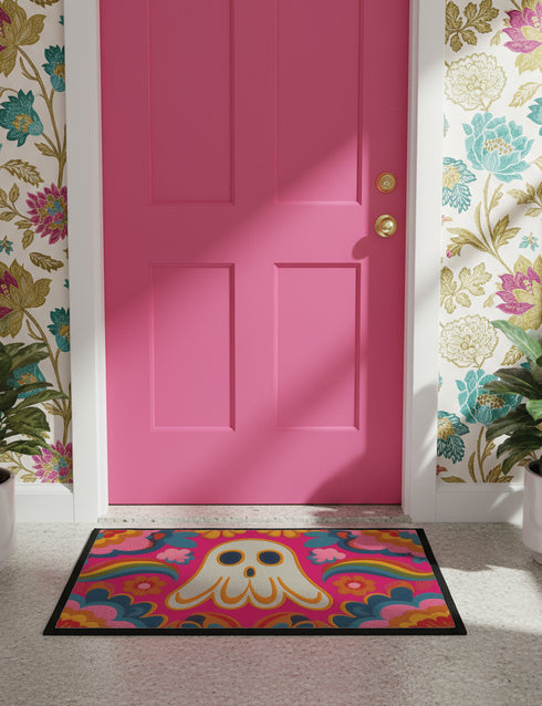A rectangular polyester doormat with a vibrant, psychedelic design on a hot pink background. In the center is a cute, cartoonish ghost in off-white with yellow and orange outlines; it has large, dark, expressive eyes. The ghost is surrounded by a maximalist pattern of 70s-style floral motifs and puffy clouds in shades of teal, blue, orange, and light pink. Two small, colorful rainbows arc over the ghost's shoulders. The doormat has a durable, textured surface and is framed by a sleek black trim.