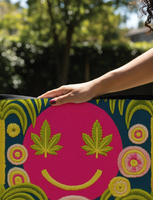 A rectangular doormat featuring a large hot pink smiley face on a deep teal background. The smiley face has green cannabis leaves for eyes and a yellow stitched-style smile. The central face is surrounded by stylized green foliage arches and concentric circles in pink, yellow, and white, resembling abstract flowers. The design mimics a vintage needlepoint or tapestry texture but is a flat print. The mat has a solid black trim border.