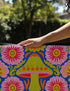 A rectangular polyester mat featuring a large central mushroom with a lime green cap, orange spots, and a pink underside. The design is surrounded by four large magenta and pink radial flowers with yellow centers. Bright green leaves and orange scrolling vines fill the spaces on a deep cobalt blue background. The artwork uses a tapestry-style visual effect to mimic the look of raised embroidery on a flat, printed surface. The mat is finished with a clean black stitched edge.
