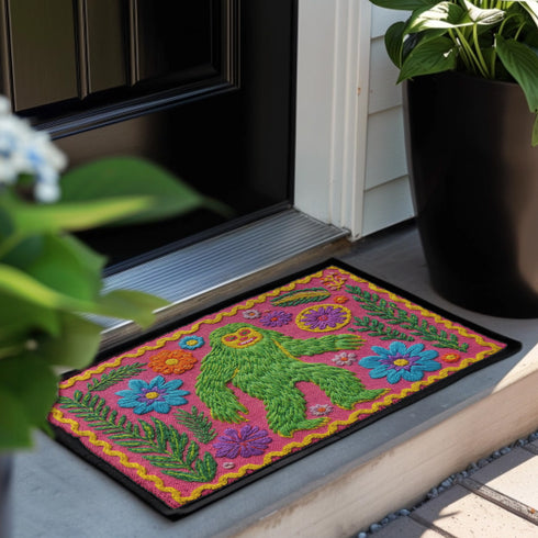 A rectangular flat-printed doormat featuring a faux-tufted, trompe-l'œil texture design. The background is a vibrant hot pink, bordered by a wavy yellow line. In the center is a bright green, smiling Bigfoot or Sasquatch figure with long shaggy hair. Surrounding the cryptid are highly detailed, colorful faux-embroidered floral motifs including blue, orange, and purple daisies, alongside green fern fronds and leaves. The mat is finished with a solid black fabric trim around the outer edge.