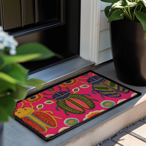 A rectangular doormat featuring a vibrant hot pink background with a high-fidelity tapestry-style print of three large, detailed beetles. The central beetle is emerald green and gold with navy accents, flanked by two side-profile beetles in ochre and deep blue. Stylized teal and yellow sunburst flowers and small sage green leaves are scattered throughout the design. The mat has a sleek black-trimmed edge and a visible flat, durable polyester texture mimicking intricate needlework.
