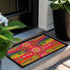 A rectangular doormat featuring a large hot pink smiley face on a deep teal background. The smiley face has green cannabis leaves for eyes and a yellow stitched-style smile. The central face is surrounded by stylized green foliage arches and concentric circles in pink, yellow, and white, resembling abstract flowers. The design mimics a vintage needlepoint or tapestry texture but is a flat print. The mat has a solid black trim border.