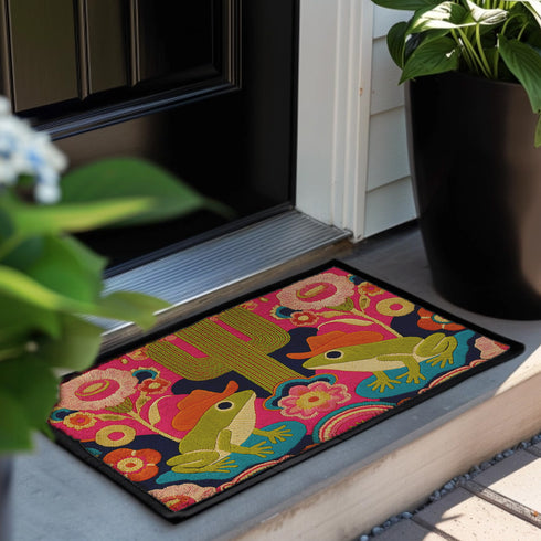 A rectangular polyester doormat with a black trim and non-slip rubber backing. The vibrant, colorful design features a symmetrical illustration on a dark navy blue background. In the center, two green frogs wearing orange cowboy hats sit facing each other. Between them is a tall, olive-green saguaro cactus. The scene is filled with large, psychedelic-style flowers in hot pink, orange, and yellow. The overall aesthetic is funky, retro 70s, maximalist, and trippy, mimicking the look of embroidery.