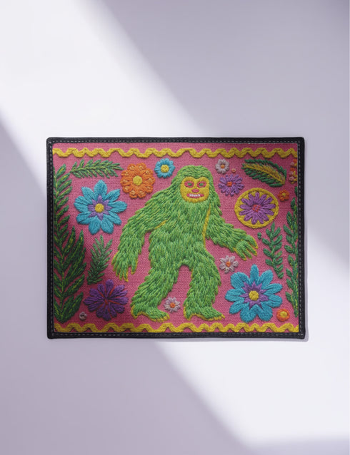 A rectangular flat-printed doormat featuring a faux-tufted, trompe-l'œil texture design. The background is a vibrant hot pink, bordered by a wavy yellow line. In the center is a bright green, smiling Bigfoot or Sasquatch figure with long shaggy hair. Surrounding the cryptid are highly detailed, colorful faux-embroidered floral motifs including blue, orange, and purple daisies, alongside green fern fronds and leaves. The mat is finished with a solid black fabric trim around the outer edge.
