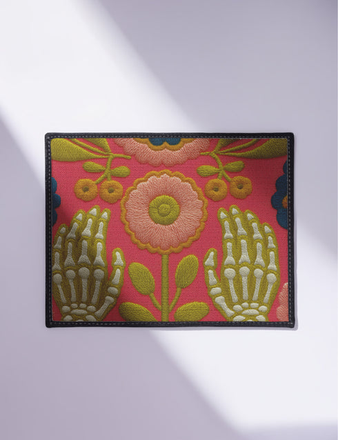 Pink outdoor doormat featuring a symmetrical folk art design. Two white skeleton hands with palms facing forward frame a large central pink and peach flower with a yellow center. Olive green stems, gold berries, and teal blue floral accents decorate the vibrant magenta background. The design mimics a textured embroidery look but is a flat 2d print. Framed with a sleek black border.