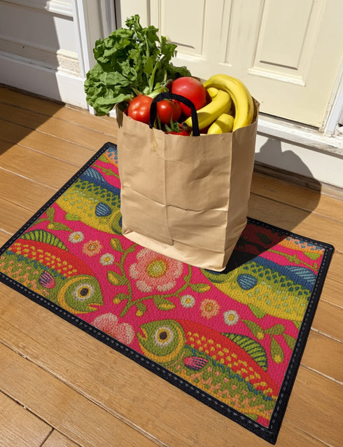 Rectangular indoor outdoor doormat featuring a vibrant hot pink background with a symmetrical maximalist pattern. The design showcases stylized sardines in yellow, lime green, and cobalt blue swimming horizontally towards a central vertical axis of blooming pale pink flowers and green vines. The imagery mimics the texture of vintage needlepoint embroidery but is a flat print on polyester. The mat is framed by a sleek black trim and features a non-slip rubber backing for safety.
