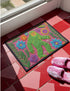 A rectangular flat-printed doormat featuring a faux-tufted, trompe-l'œil texture design. The background is a vibrant hot pink, bordered by a wavy yellow line. In the center is a bright green, smiling Bigfoot or Sasquatch figure with long shaggy hair. Surrounding the cryptid are highly detailed, colorful faux-embroidered floral motifs including blue, orange, and purple daisies, alongside green fern fronds and leaves. The mat is finished with a solid black fabric trim around the outer edge.