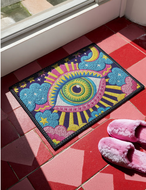 Rectangular entryway doormat with a deep purple background printed in a 2D faux embroidery illusion. The symmetrical mandala centers on a large green, pink, and white evil eye with golden yellow sunburst lines radiating outward. The corners hold textured cyan clouds, yellow crescent moons, and stars, with a scalloped floral motif along the lower edge. Finished with black trim and a non-slip rubber backing. Psychedelic celestial maximalist home decor and protective evil eye welcome mat for bold, eclectic ent