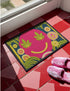 A rectangular doormat featuring a large hot pink smiley face on a deep teal background. The smiley face has green cannabis leaves for eyes and a yellow stitched-style smile. The central face is surrounded by stylized green foliage arches and concentric circles in pink, yellow, and white, resembling abstract flowers. The design mimics a vintage needlepoint or tapestry texture but is a flat print. The mat has a solid black trim border.