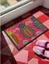 A rectangular doormat featuring a vibrant hot pink background with a high-fidelity tapestry-style print of three large, detailed beetles. The central beetle is emerald green and gold with navy accents, flanked by two side-profile beetles in ochre and deep blue. Stylized teal and yellow sunburst flowers and small sage green leaves are scattered throughout the design. The mat has a sleek black-trimmed edge and a visible flat, durable polyester texture mimicking intricate needlework.