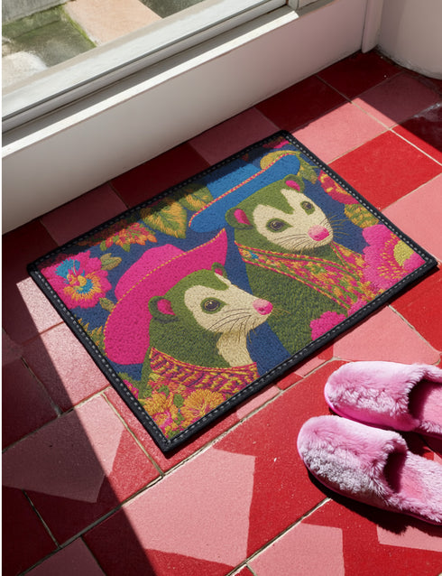 A rectangular non-slip doormat with a black trimmed edge. The mat features a vibrant, colorful folk art illustration on a dark navy blue background with a faux embroidered texture. The design depicts two friendly opossums, rendered in an olive green color with white faces and pink noses. They are surrounded by large, stylized flowers and leaves in shades of hot pink, red, turquoise, and yellow, creating a busy, maximalist pattern. The overall aesthetic is eclectic, whimsical, & highly detailed.