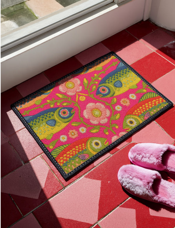 Rectangular indoor outdoor doormat featuring a vibrant hot pink background with a symmetrical maximalist pattern. The design showcases stylized sardines in yellow, lime green, and cobalt blue swimming horizontally towards a central vertical axis of blooming pale pink flowers and green vines. The imagery mimics the texture of vintage needlepoint embroidery but is a flat print on polyester. The mat is framed by a sleek black trim and features a non-slip rubber backing for safety.