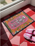 A rectangular terracotta orange doormat featuring a central illustration of a white skeleton lying inside a pink coffin shape. The design is surrounded by a symmetrical pattern of green leaves, pink flowers, and red and white spotted mushrooms. The imagery uses a trompe-l'œil effect to mimic the texture of raised needlepoint embroidery, but the surface is flat printed polyester. The mat is framed with a solid black edge.
