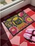 High-fidelity printed doormat featuring a "Space Kitty" design in a tapestry-style faux-texture. A black cat with large yellow eyes wears a golden ribbed space helmet, centered on a deep navy background. The cat is surrounded by oversized, vibrant magenta and pink folk-art flowers with chartreuse and olive green leaves. The design uses a printed illusion to mimic 3D embroidery stitching. The mat is rectangular with a sleek black border and a non-slip backing.