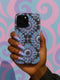 A phone case featuring a repetitive geometric pattern of iridescent floral eyeballs with a glossy finish. The case has a two-piece design constructed from a tough, black polycarbonate shell and a flexible TPU liner for superior impact resistance. Each circular eyeball motif is centered within a four-panel grid, with the flower petals fanning out in a soft, holographic blue, purple, and green sheen that shifts with the light. The black background of the case provides a dark, goth aesthetic.