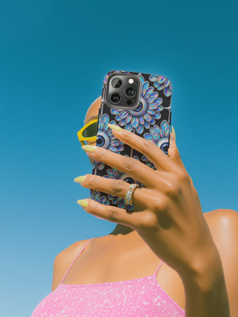 A phone case featuring a repetitive geometric pattern of iridescent floral eyeballs with a glossy finish. The case has a two-piece design constructed from a tough, black polycarbonate shell and a flexible TPU liner for superior impact resistance. Each circular eyeball motif is centered within a four-panel grid, with the flower petals fanning out in a soft, holographic blue, purple, and green sheen that shifts with the light. The black background of the case provides a dark, goth aesthetic.