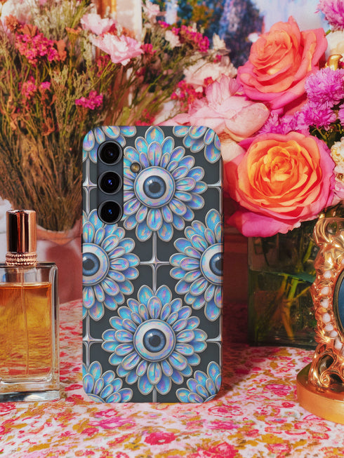 A phone case featuring a repetitive geometric pattern of iridescent floral eyeballs with a glossy finish. The case has a two-piece design constructed from a tough, black polycarbonate shell and a flexible TPU liner for superior impact resistance. Each circular eyeball motif is centered within a four-panel grid, with the flower petals fanning out in a soft, holographic blue, purple, and green sheen that shifts with the light. The black background of the case provides a dark, goth aesthetic.