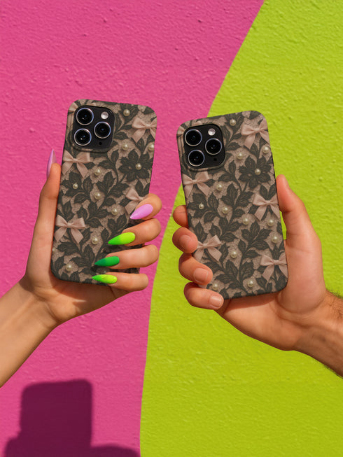 Phone case with black lace floral design, soft pink satin bows, and small pearl accents on a pale nude background; fits iPhone, Samsung, and Pixel models