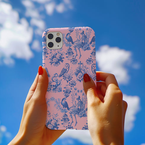 Phone case with pink background featuring blue toile print of skeletons, peacocks, owls, and roses. Victorian-inspired design with gothic elements. Dual-layer protection for various smartphone models.