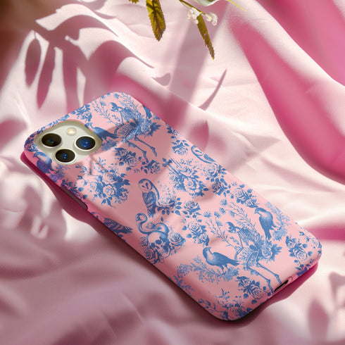 Phone case with pink background featuring blue toile print of skeletons, peacocks, owls, and roses. Victorian-inspired design with gothic elements. Dual-layer protection for various smartphone models.