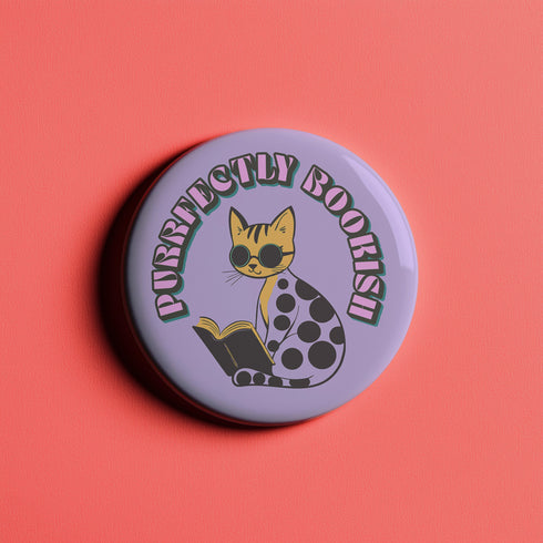 Purple button pin featuring a yellow cat wearing sunglasses and reading a book, with "Purrfectly Bookish" text curved around the edge