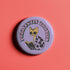 Purple button pin featuring a yellow cat wearing sunglasses and reading a book, with "Purrfectly Bookish" text curved around the edge