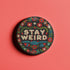 Stay Weird" black pinback button with pearl-effect lettering surrounded by folk art flowers in red and turquoise on a dark background.